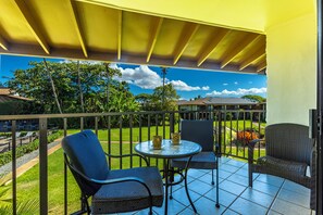 A balcony with two wicker chairs and a small table, overlooking a well-maintained garden and a clear blue sky.