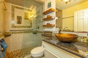 A bathroom with a glass shower enclosure, a white toilet, a large vessel sink, and wooden shelves above the sink.