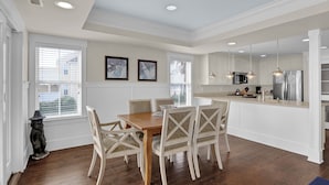 A dining area with a wooden table and chairs, a kitchen with white cabinets and stainless steel appliances, and framed artwork on the wall. - C Street A - Incredible Ocean Front Condo in the heart of Wrightsville Beach (Wrightsville Beach)