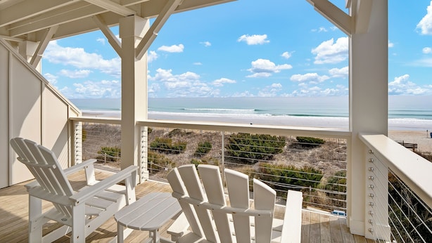 A covered outdoor seating area with white chairs and a table, overlooking a beach with the ocean and sky. - C Street A - Incredible Ocean Front Condo in the heart of Wrightsville Beach (Wrightsville Beach)