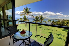 Ein Balkon mit Glastisch und Stühlen, mit Blick auf eine Rasenfläche, Palmen und den Ozean.