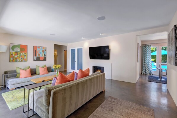 A modern living room with a sofa, a coffee table, and a fireplace. There are abstract paintings on the walls and a television mounted above the fireplace. - Palmera - Tahquitz River Estates Private Paradise! (Palm Springs)