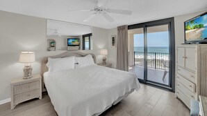 Ein Schlafzimmer mit Bett, Deckenventilator, Fernseher, Nachttisch, Kommode und einem Balkon mit Blick auf den Strand.
