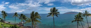 A tropical beach with clear turquoise waters, palm trees, and a distant mountain.