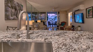 Quarto de hotel moderno com bancada de granito, pia de aço inoxidável, TV de tela plana e uma grande janela com vista para palmeiras.