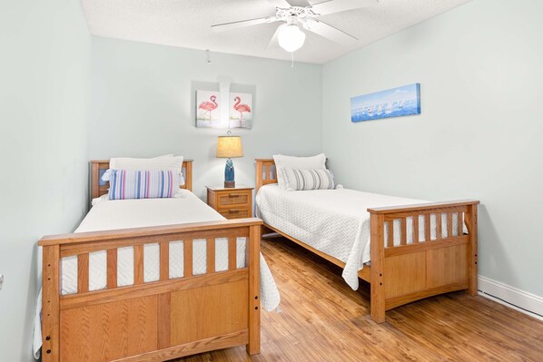 A bedroom with two wooden beds, a ceiling fan, a nightstand with a lamp, and wall art. - Sandpiper 303 | Beach Views, Steps from Ocean, Prime South End Location! (Tybee Island)