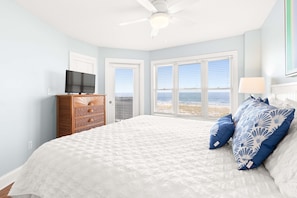 Ein Schlafzimmer mit einem großen Bett, einem Fernseher auf einer Kommode und Blick auf den Strand durch die Fenster.