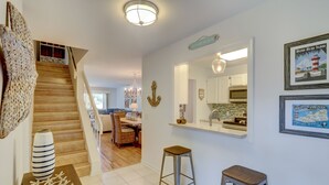 A modern kitchen with white cabinets, a built-in oven, and a microwave. There is a dining area with wicker chairs and a chandelier.