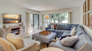 A spacious living room with a large sofa, a television, and a view of the outdoors through large windows.