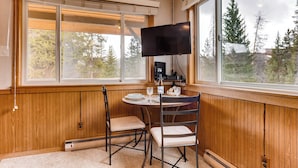 A room with a wooden paneled wall, a large window with a view of trees, a table with chairs, a flat-screen TV mounted on the wall, and a thermostat on the floor. - Cozy Top Floor Unit w/ Lots of Windows. Ski In, Steps from Main Street | PM8D (Breckenridge)