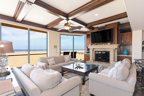 A spacious living room with a large TV, comfortable seating, a fireplace, and a view of the beach. - 3425O Oceanfront Tuscan Villa (Oxnard)
