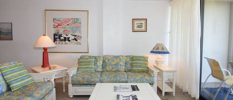 Un salón con un sofá estampado con motivos florales, una mesa de centro blanca con revistas, una lámpara roja y una lámpara azul sobre una mesita auxiliar.
