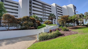 A modern multi-story hotel with a white facade, multiple balconies, and landscaped gardens with various shrubs and trees.