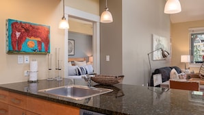 A modern kitchen with granite countertops, a built-in dishwasher, and a large sink. There is a colorful abstract painting on the wall, a woven basket on the counter, and a cozy seating area with a sofa and a coffee table.