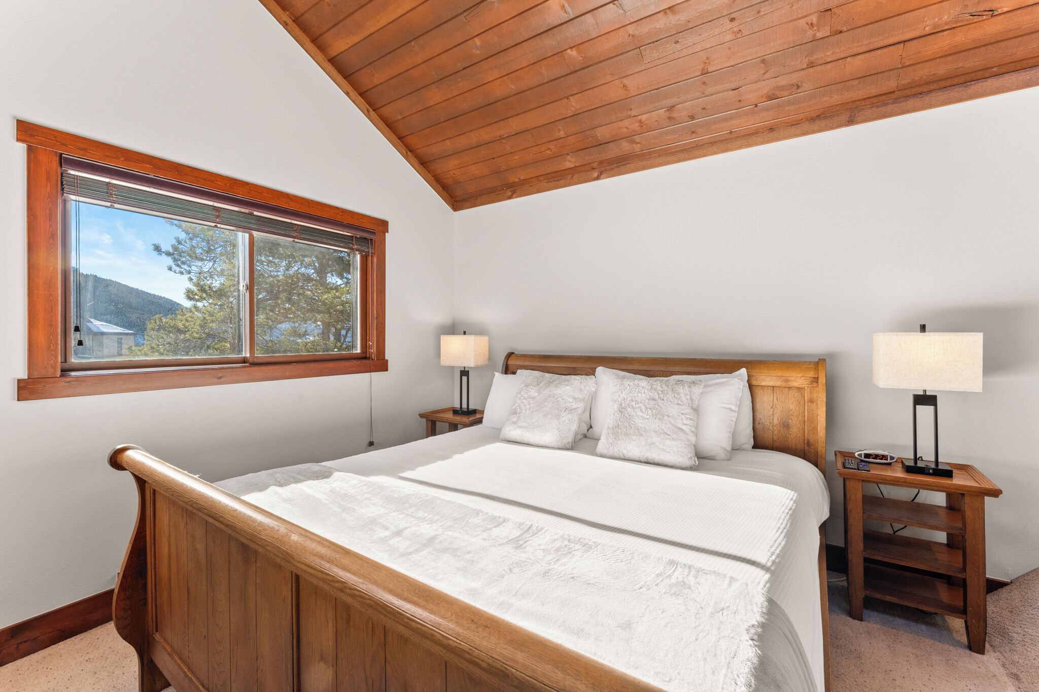 A bedroom with a wooden bed, two bedside tables with lamps, a window with a view of trees and a house, and a sloped ceiling.