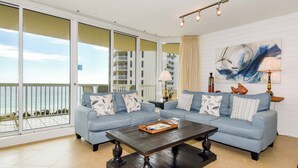 A living room with a blue sofa set, a wooden coffee table, and a large window with a view of a building and a balcony. - Spacious beachfront end condo w/large private balcony! 🌴 (Destin)