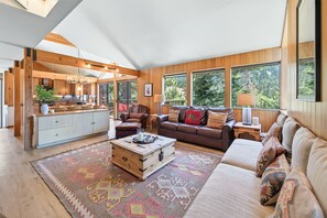 A spacious living area with a wooden floor, a large sofa, a coffee table, and a kitchen area with wooden cabinets and a dining table. - Juniper at Alpine - Private Hot tub, Pet Friendly & Amazing Views! (Alpine Meadows)