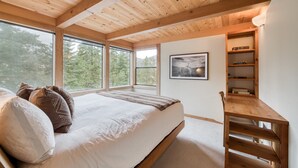 Ein Schlafzimmer mit einem großen Bett, Holzbalkendecke, einer eingebauten Bücherwand und großen Fenstern mit Blick auf Bäume.