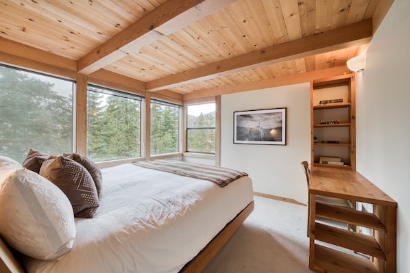 A bedroom with a large bed, wooden ceiling beams, a built-in bookshelf, and large windows offering a view of trees.