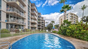 A swimming pool surrounded by multi-story buildings with balconies, palm trees, and a clear sky.