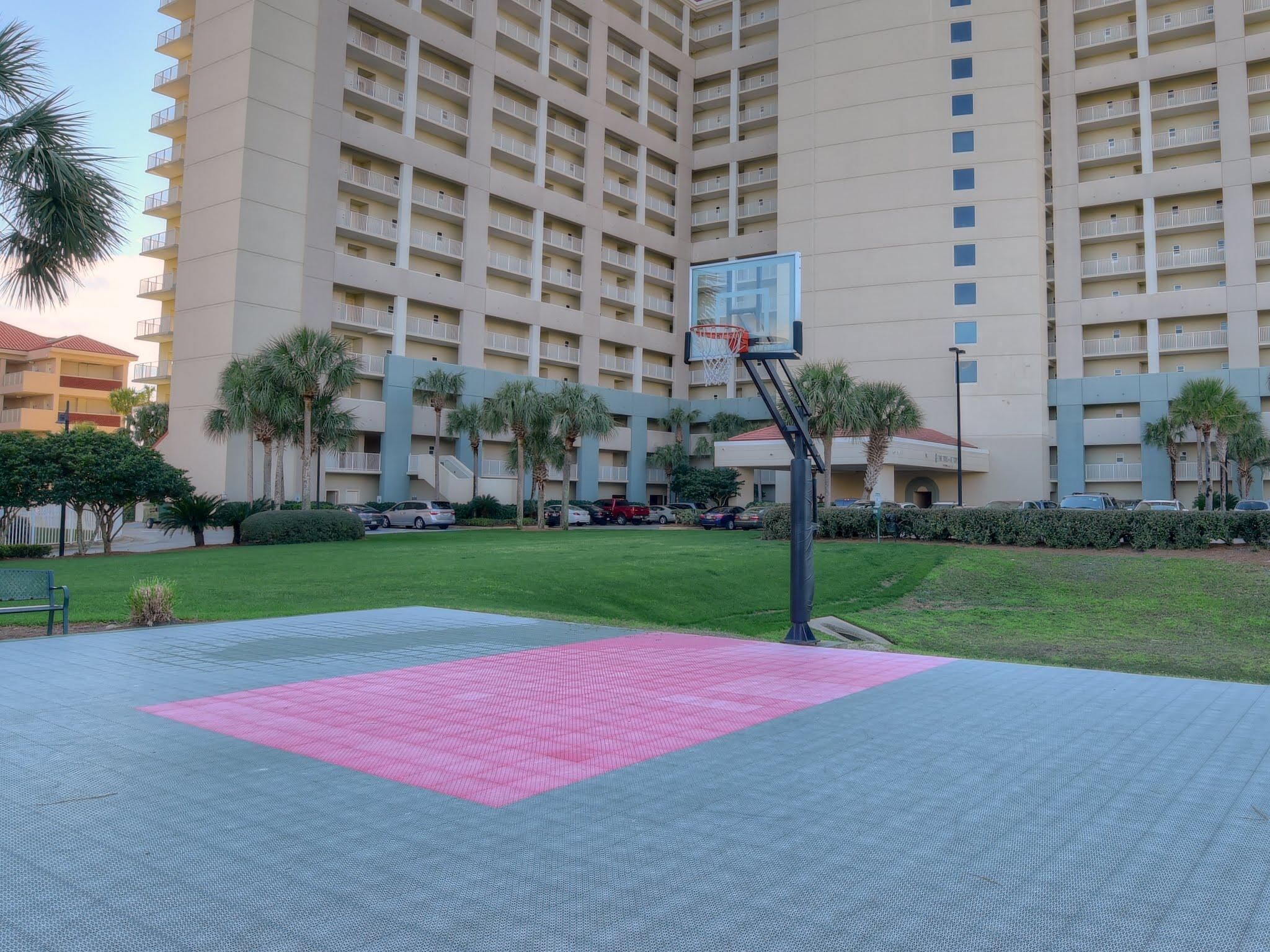 Un terrain de basket avec un tapis rose au centre, entouré d’une pelouse verte et de palmiers, et un grand immeuble en arrière-plan.