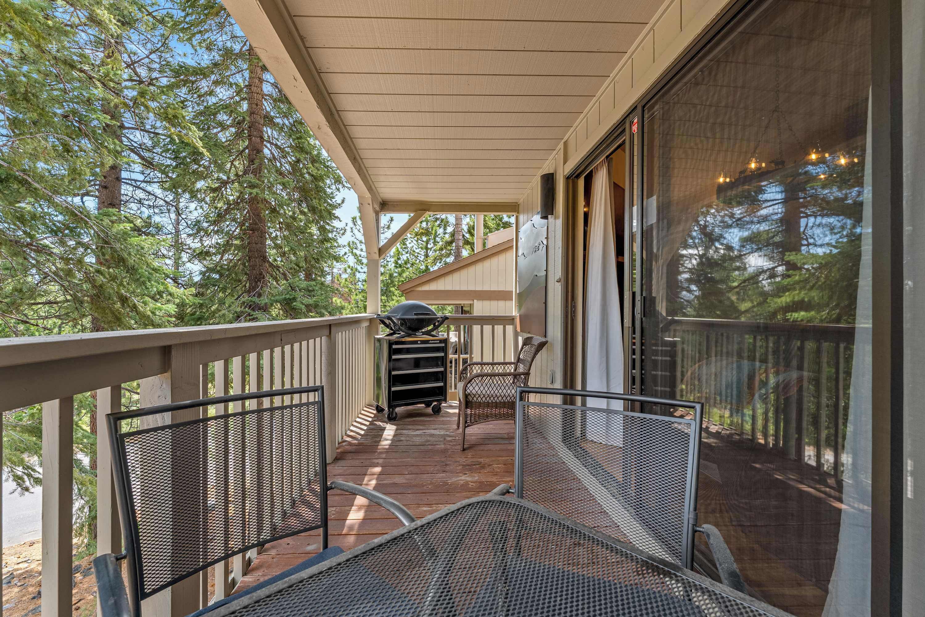A covered outdoor deck with a grill, chairs, and a view of trees.