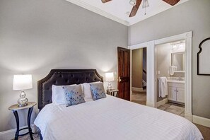 A bedroom with a large bed, two bedside tables with lamps, a ceiling fan, and a view into the bathroom.