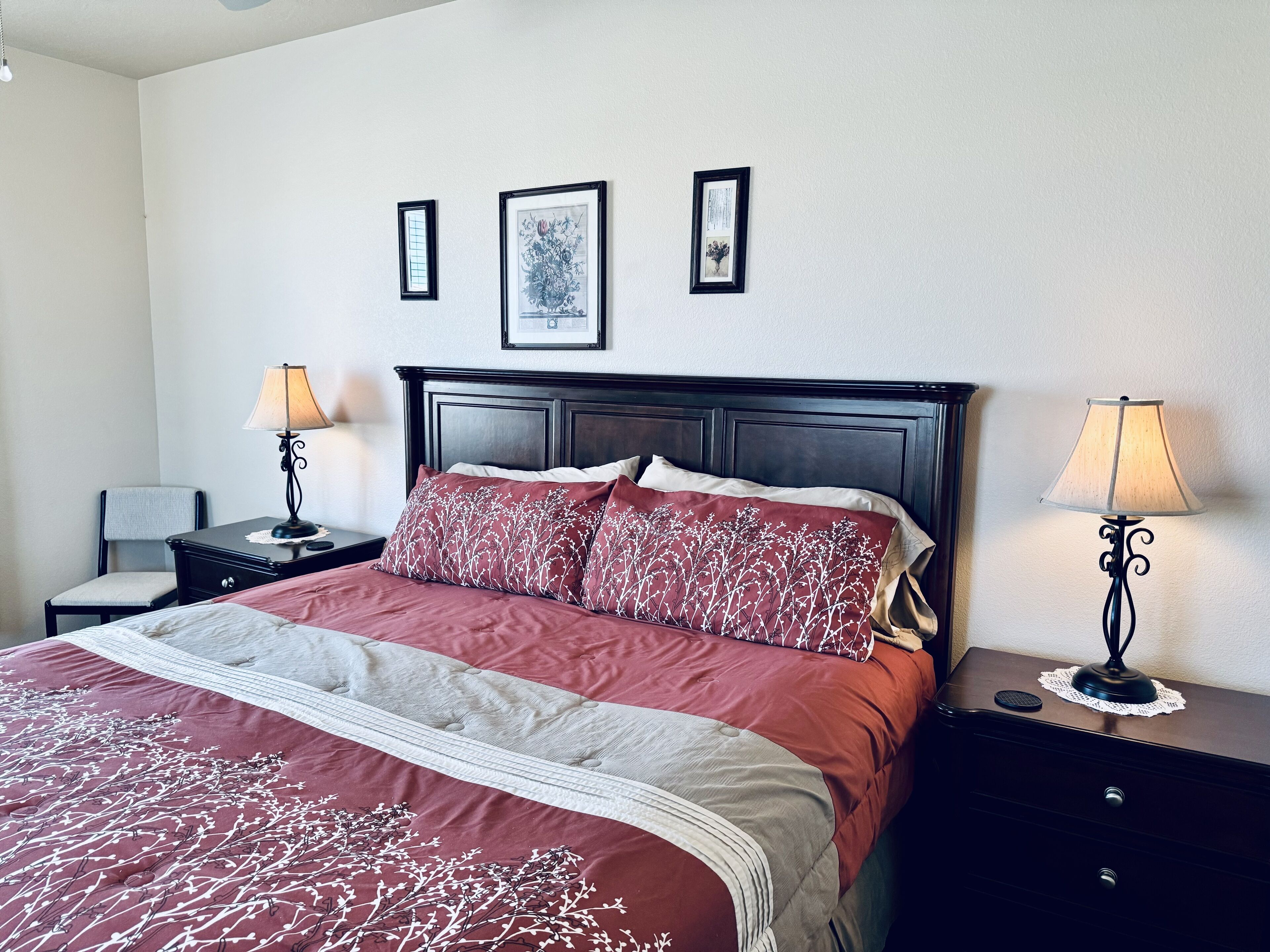 Un lit bien fait, recouvert d’une couvre-lit à motifs rouges et blancs, est encadré de deux tables de chevet sur lesquelles reposent des lampes. On y voit également une chaise et des tableaux encadrés accrochés au mur.