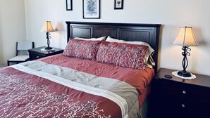 Ein ordentlich bezogenes Bett mit einer rot-weißen Bettdecke, flankiert von zwei Nachttischen mit Lampen, einem Sessel und gerahmten Bildern an der Wand.