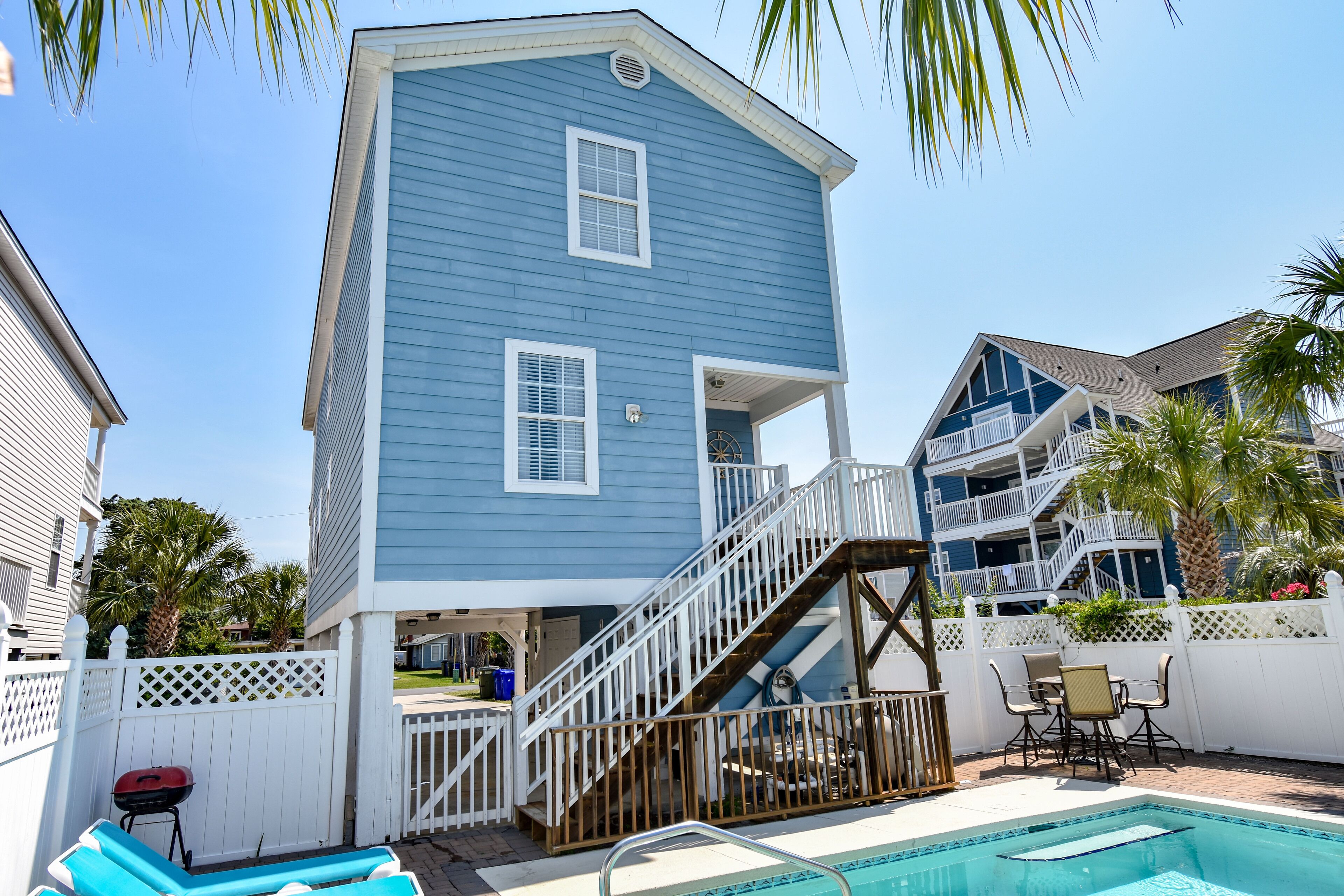 A two-story house with a blue exterior, a wooden staircase leading to a balcony, a pool, and a patio area with outdoor furniture.