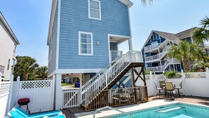 Una casa de dos pisos con fachada azul, una escalera de madera que conduce a un balcón, una piscina y un patio con mobiliario exterior.