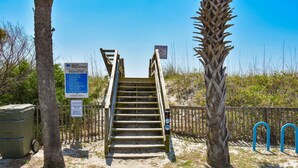 Una escalera de madera que conduce a una zona de playa, con un letrero que dice 'Niños Perdidos', un letrero que indica 'No se permiten perros' y un contenedor de basura.
