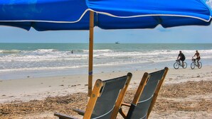 Une plage avec un parasol bleu, deux chaises en bois et une personne à vélo.