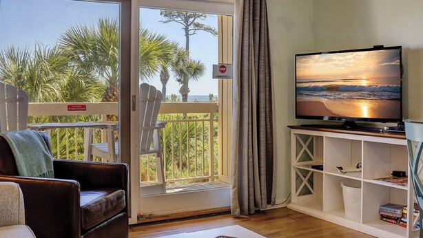 Un salon avec un téléviseur diffusant une image de coucher de soleil, un balcon avec un panneau de signalisation indiquant un arrêt, et une étagère blanche remplie de livres.