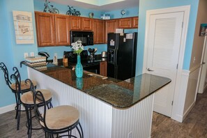 A kitchen with wooden cabinets, a black refrigerator, a microwave, and a granite countertop. There are two chairs and a vase with flowers on the countertop.