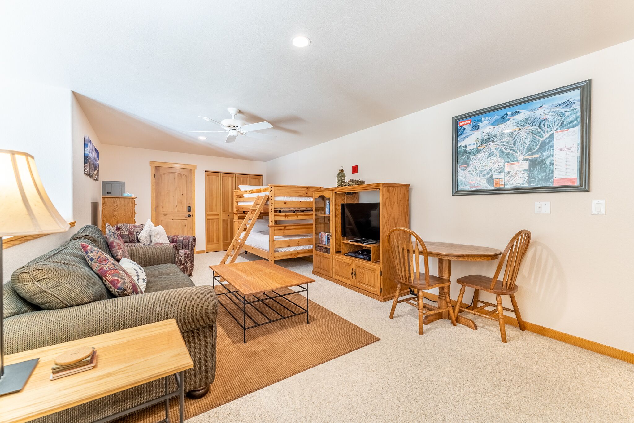 A cozy living room with a sofa, coffee table, and a dining area with chairs. There is a wooden cabinet with a TV and a ceiling fan.