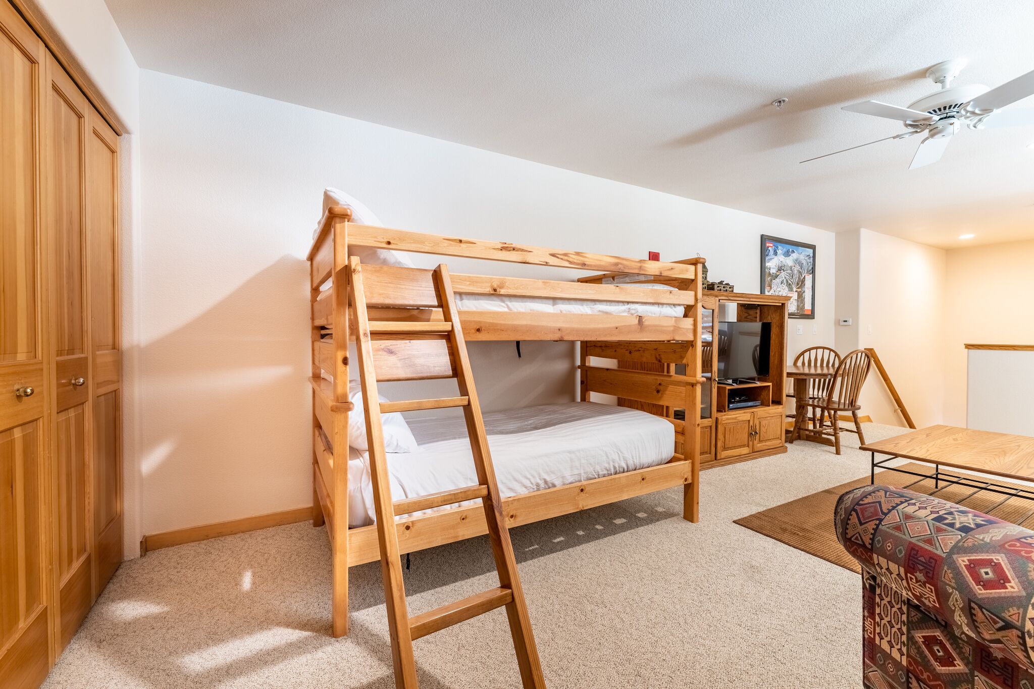Een stapelbed met een ladder, een houten kast, een televisie, een eettafel en een gestreept sofa in een kamer met witte muren en een plafondventilator.