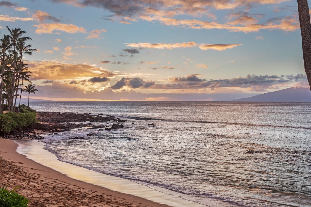 Una playa con costa rocosa, palmeras y una puesta de sol sobre el océano.