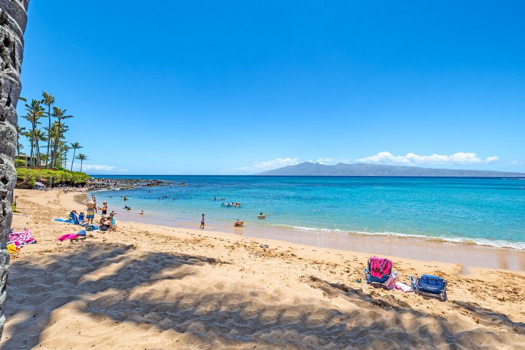 Une plage avec une eau d’un bleu limpide, des gens qui se baignent et qui prennent des bains de soleil, et une montagne en arrière-plan.