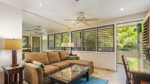 A living room with a sofa, coffee table, and a dining table with chairs. There is a ceiling fan, a lamp, and a large window with blinds. - Aloha Condos, Napili Bay Resort, Condo 217, Beach View (Lahaina)
