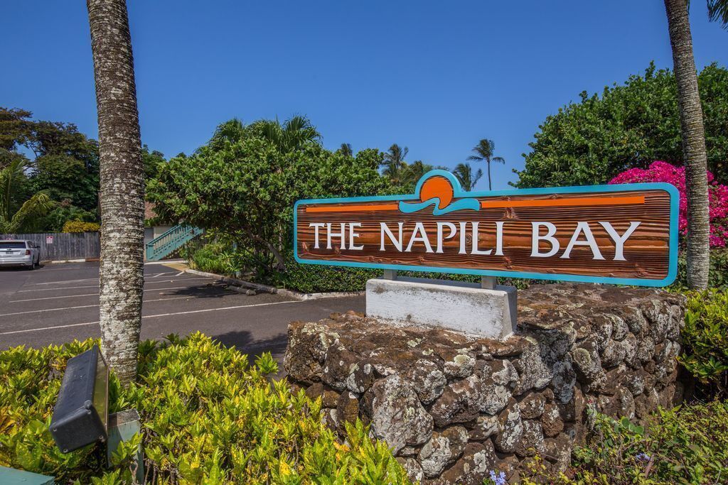 A wooden sign with 'THE NAPI L BAY' written on it, surrounded by greenery and a clear sky.