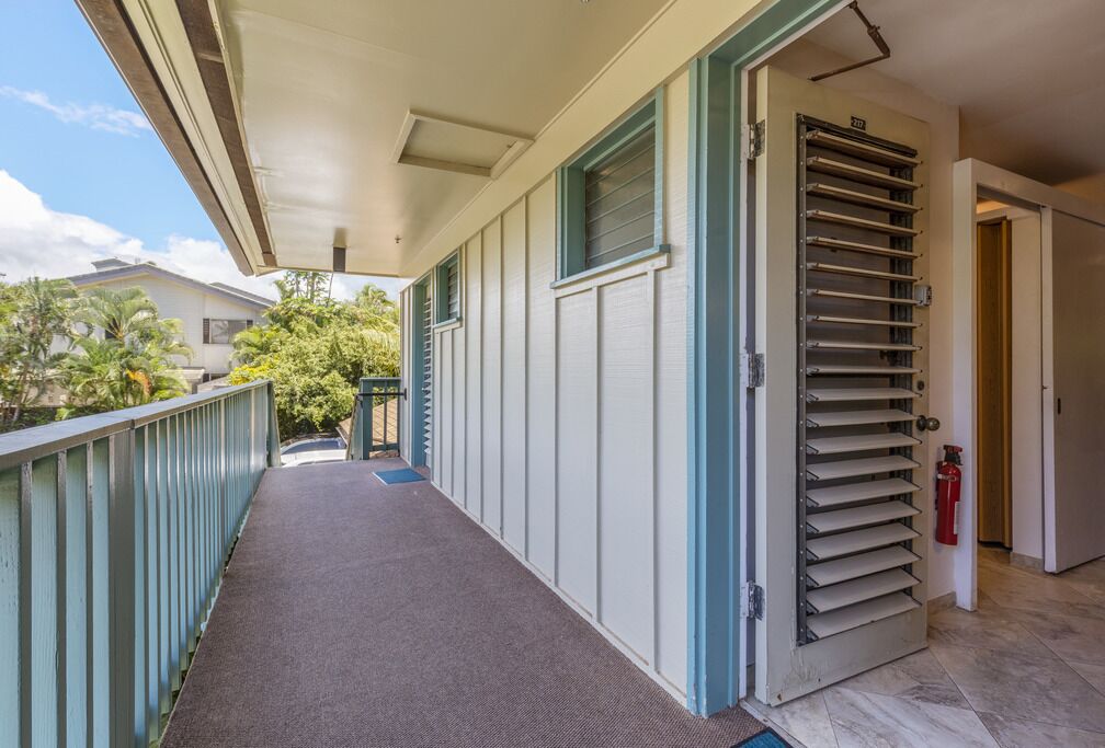 A covered outdoor corridor with a metal door, a fire extinguisher, and a view of a residential area with palm trees.