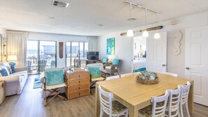Un vaste espace de vie comprenant une table à manger en bois, des chaises blanches et des meubles en osier. Un téléviseur est fixé sur un meuble en bois, et une grande fenêtre offre une vue sur l’extérieur.