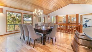A spacious dining area with a wooden ceiling, large windows offering a view of trees and mountains, and a modern chandelier.