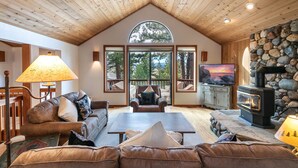 A cozy living room with a stone fireplace, leather sofas, a television, and large windows offering a view of trees and mountains.