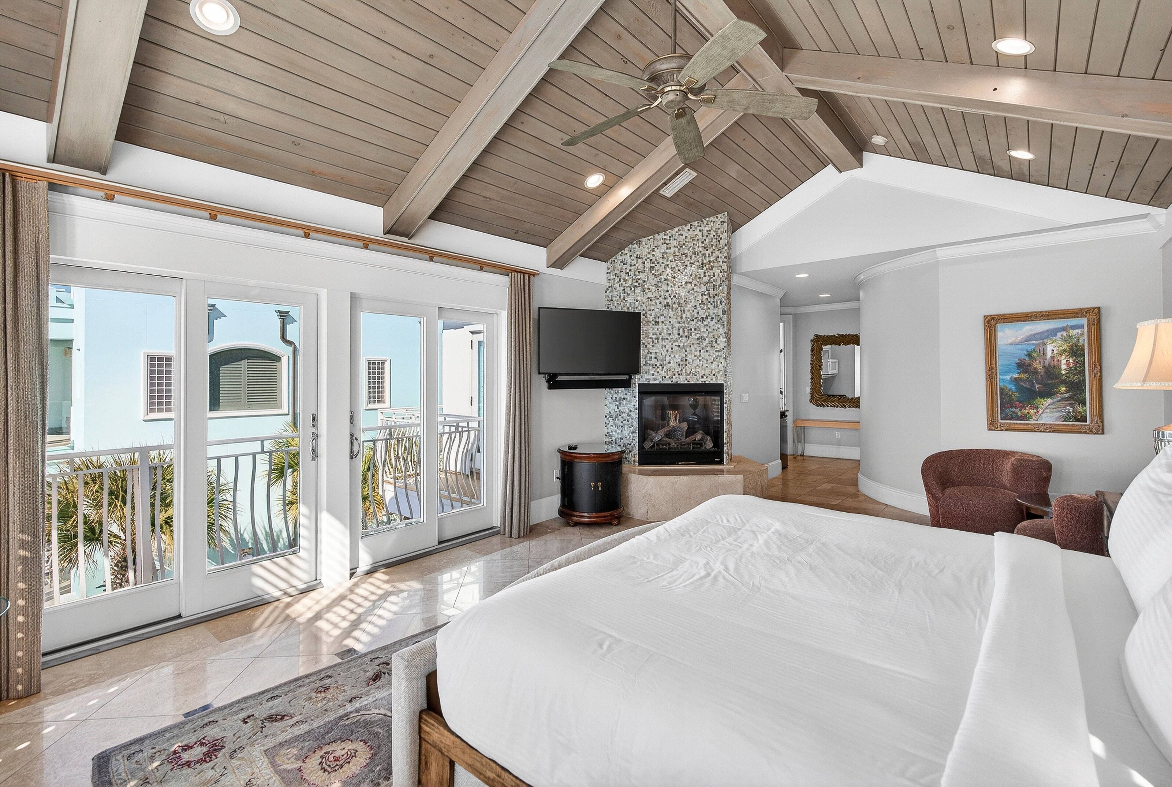 A spacious bedroom with a large bed, a television, a fireplace, and a balcony with a view of the outside.