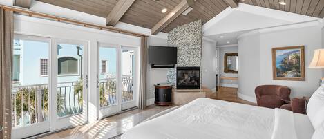 A spacious bedroom with a large bed, a television, a fireplace, and a balcony with a view of the outside.
