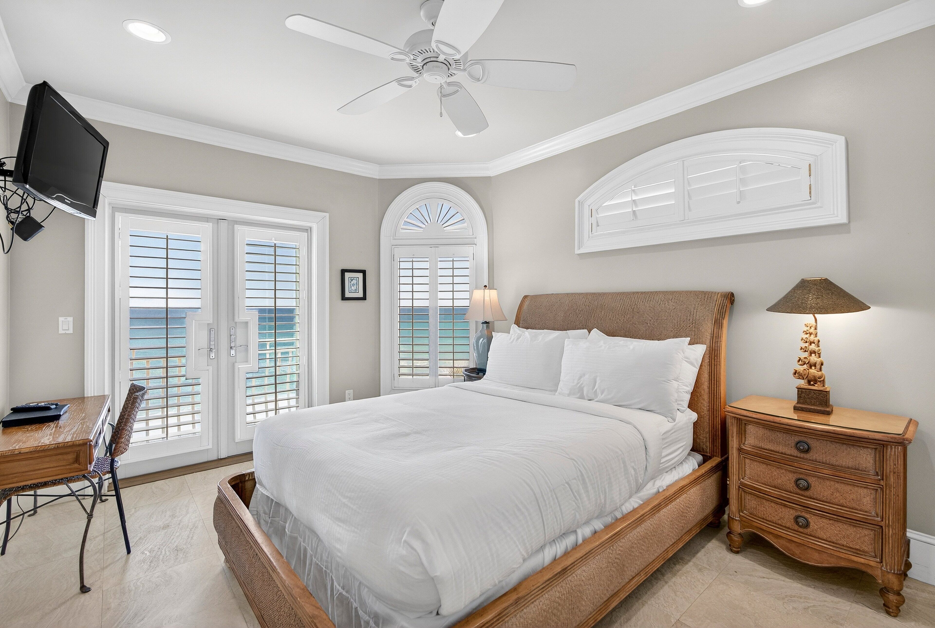 A bedroom with a large bed, a desk, a chair, a nightstand with a lamp, and a ceiling fan.