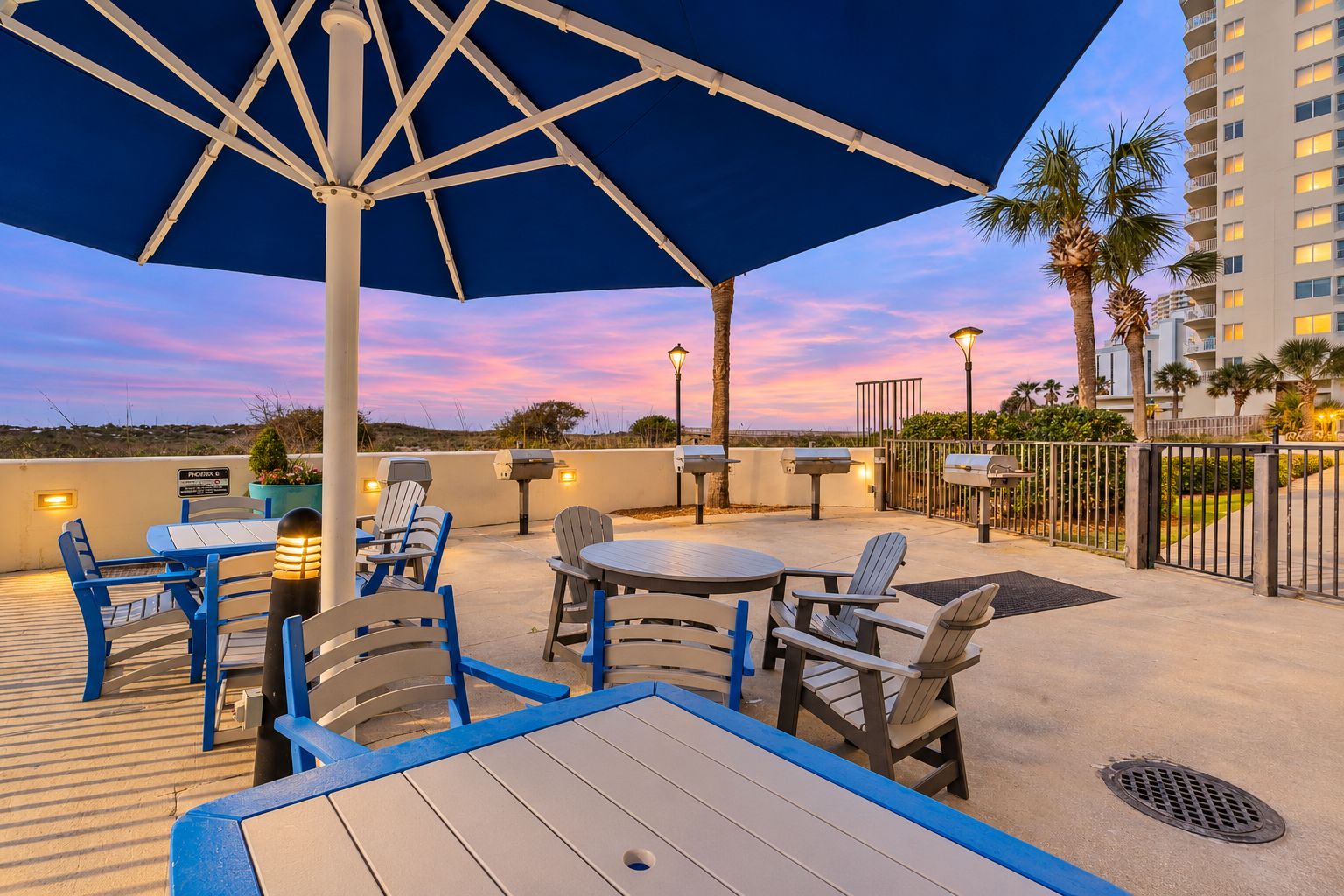 An outdoor patio with tables and chairs, a large umbrella, and a view of a building in the background.