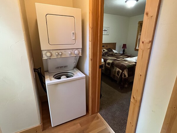 Un lavadero pequeño con lavadora y secadora, piso de madera y una puerta que da a un dormitorio con cama y mesita de noche.
